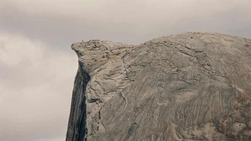 The video shows a closeup view of a massive imposing rock formation likely the iconic half dome under a cloudy sky small figures of people are visible on the very top edge of the peak highlighting its immense scale