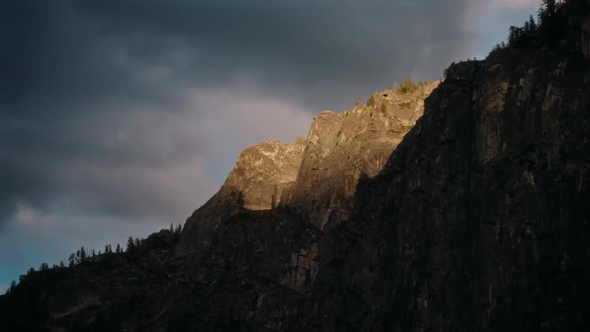 Storm clouds roll over a rocky mountain with trees dotting the top