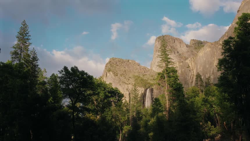 A waterfall cascades down rocky cliffs surrounded by lush green trees beneath a partly cloudy sky