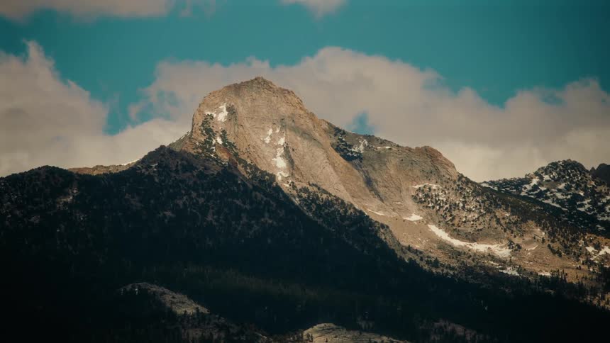 The video displays a grand shot of a rugged mountain peak partially illuminated by sunlight rising prominently against a sky with fluffy clouds the lower slopes of the mountain are covered with dark green trees and patches of snow are visible near the