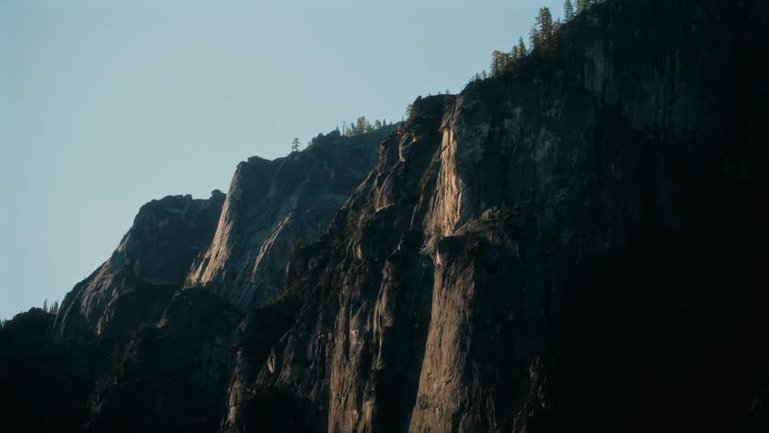 Sunlit cliffs with rugged rock formations rise sharply beneath a clear sky with sparse trees lining the upper edges