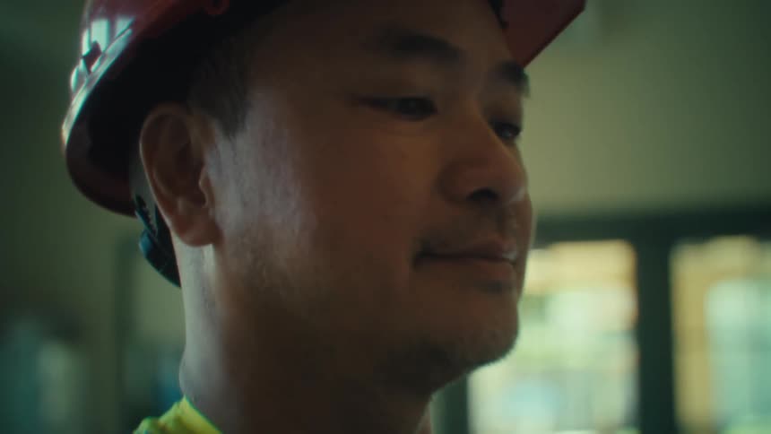 A construction worker indoors wearing a helmet and safety vest looking at an invoice with a smile on his face