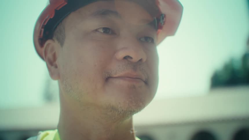 Shot of an east asian man likely in his 40s or 50s wearing an orange hard hat and a yellow safety vest over a black tshirt he is looking down intently at a white tablet