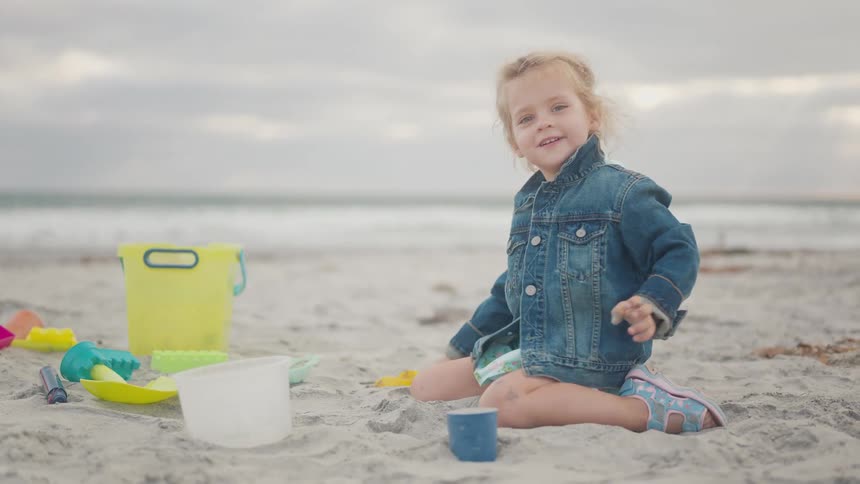 A young child with blonde curly hair wearing a denim jacket and patterned shorts is playing in the sand on a beach they are surrounded by various colorful sand toys and the ocean is visible in the background under a cloudy sky