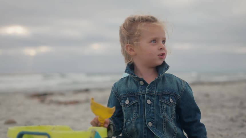 A young child with blonde hair dressed in a denim jacket is playing on a sandy beach they hold a yellow sand shovel and look around with an expressive face occasionally touching their head or hair