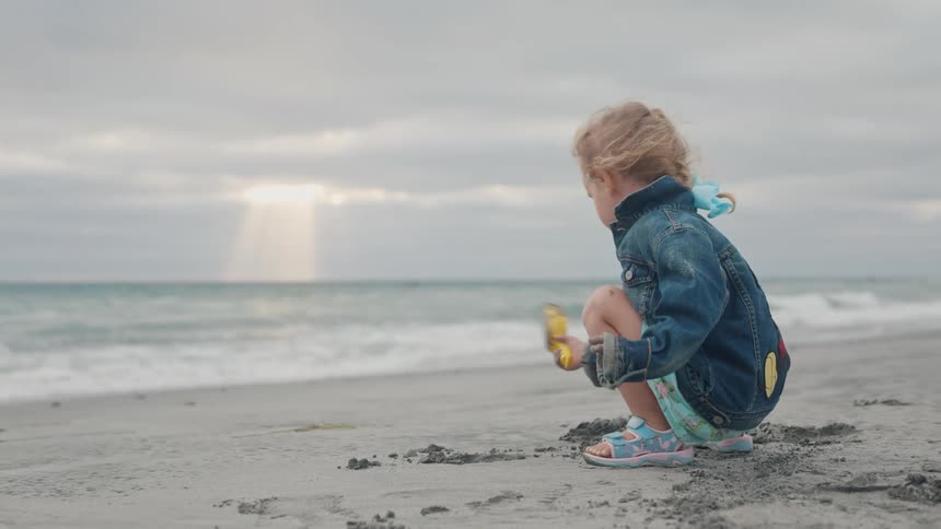 A child in a blue jacket and light dress plays on a sandy beach with a yellow shovel as sunlight filters through soft clouds during sunset