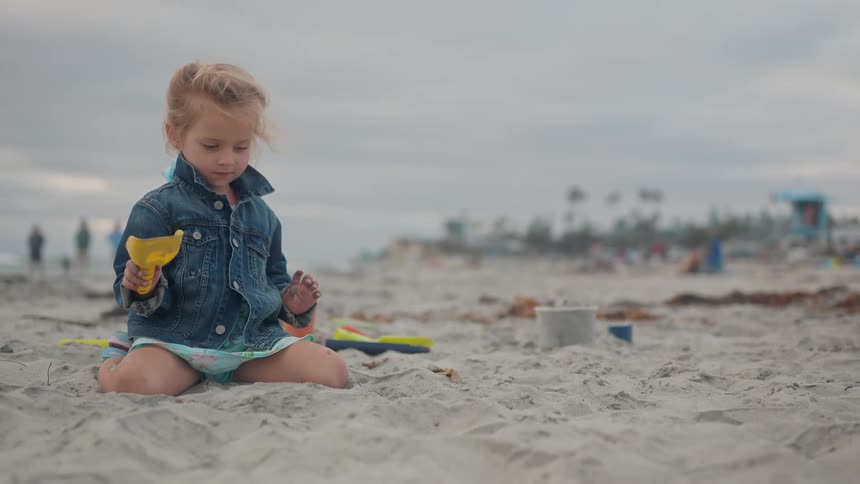 A small child in a denim jacket and patterned shorts kneels on a sandy beach playing with a yellow sand toy while looking around at the blurred background of people and distant structures