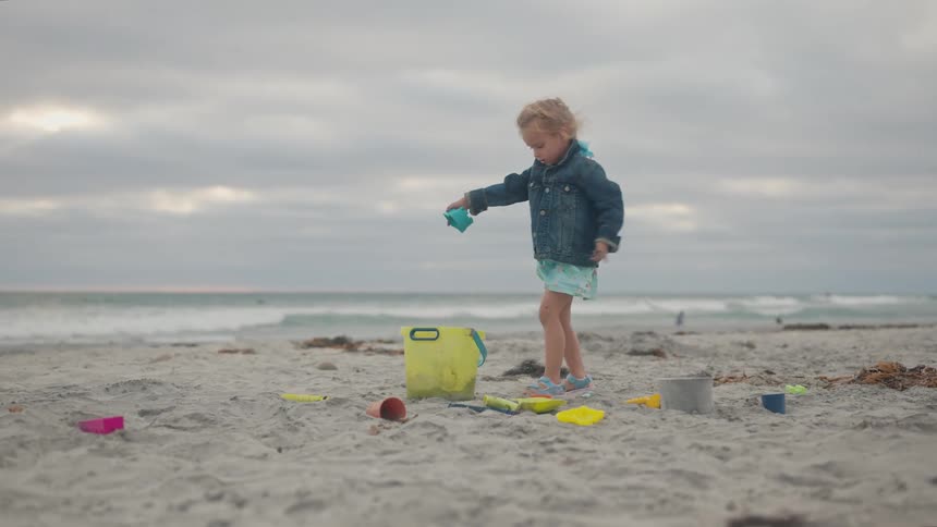 A child in a blue jacket and light dress plays on a sandy beach pouring sand or water from a blue container into a yellow bucket surrounded by scattered beach toys