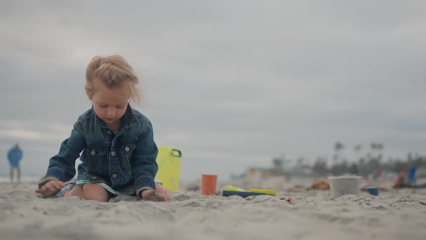 A child in a blue jacket and light dress plays on a sandy beach surrounded by scattered beach toys