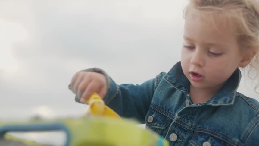 A child in a blue jacket and light dress plays on a sandy beach with a yellow shovel beneath soft clouds at sunset
