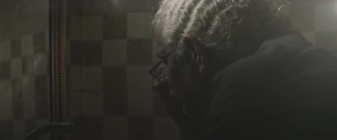 An older man taking off his glasses in a bathroom as he looks in the mirror