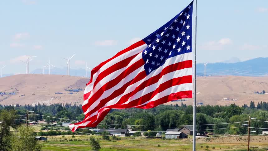 An american flag flying on a pole in the wind with a power line in front a small town beyond and a windmill in the background at midday