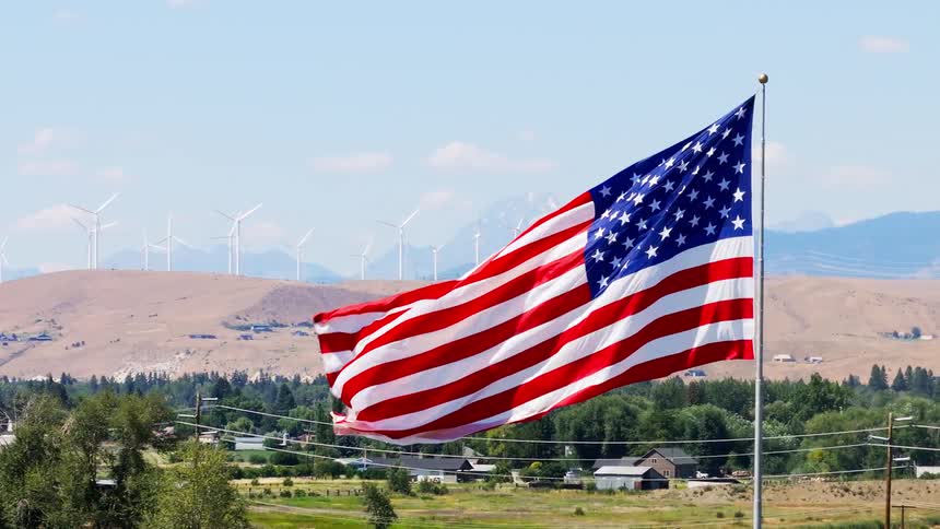 Us flag in the foreground with some urban homes in the background and wind turbines
