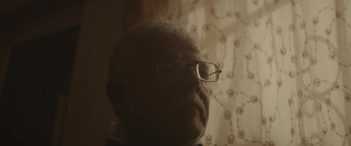 An elderly man sits in a room next to curtains