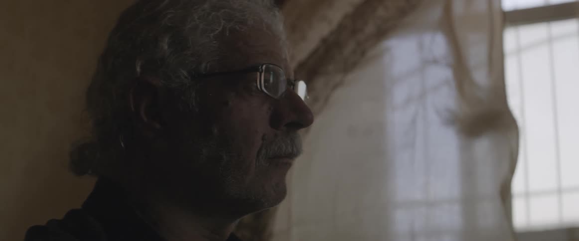 An elderly man looks out a window as curtains blow in the breeze