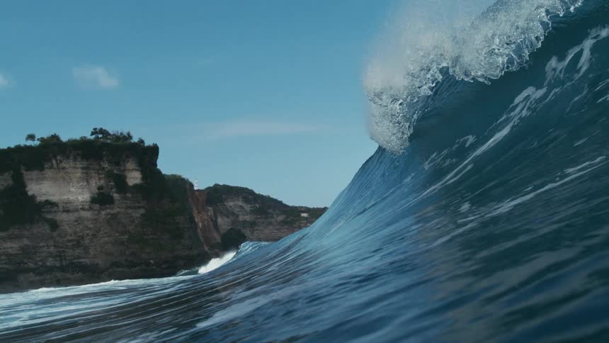 A powerful ocean wave rises near rocky coastal cliffs under a clear blue sky