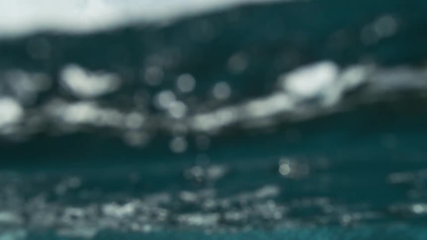 A powerful ocean wave crests and foams beneath a partly cloudy sky with water droplets suspended midair rolling over the camera