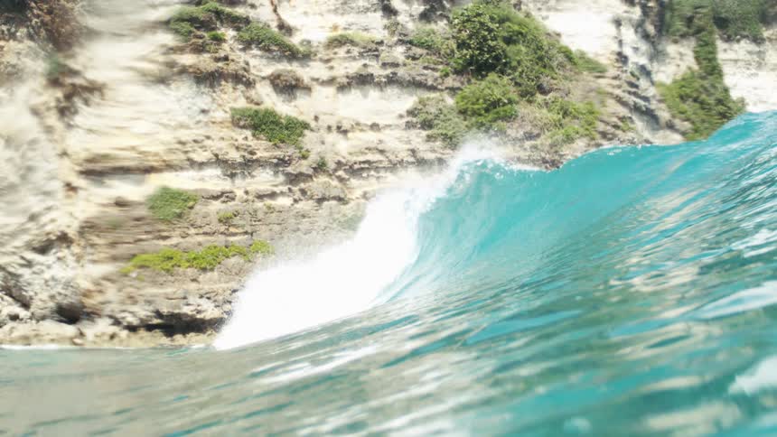A wave crests in the ocean near a rocky cliff before the camera goes underwater