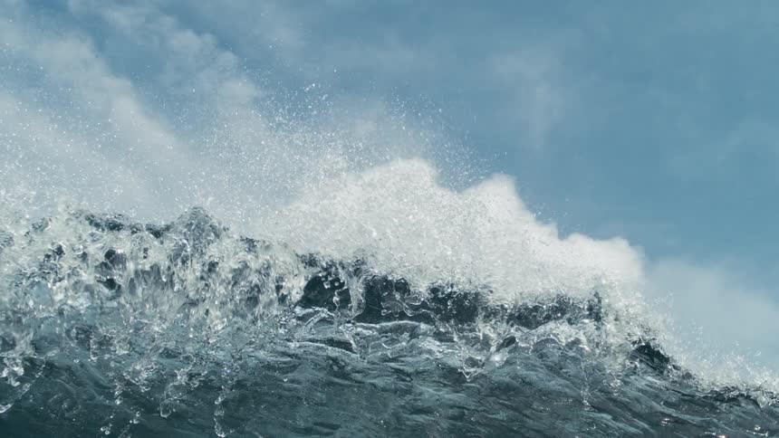 A deep blue ocean wave with textured surface is captured against a cloudy sky