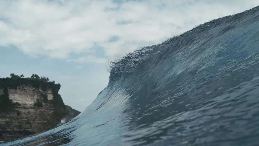 A large wave curls and breaks against rocky cliffs topped with green vegetation beneath a partly cloudy sky
