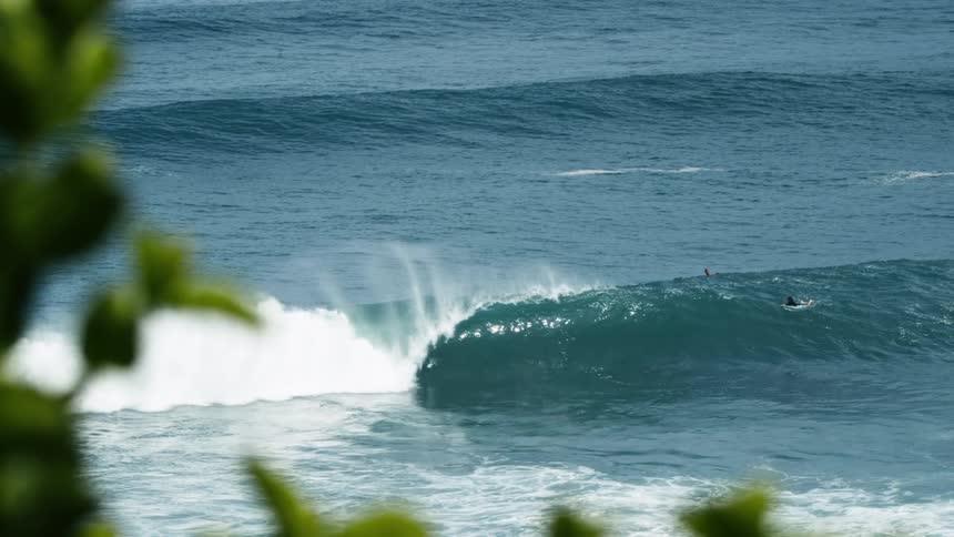 Filmed in slow motion from a leafy coastline a skilled surfer catches a large perfectly formed ocean wave and gets a long barrel ride disappearing deep inside the tube as it breaks