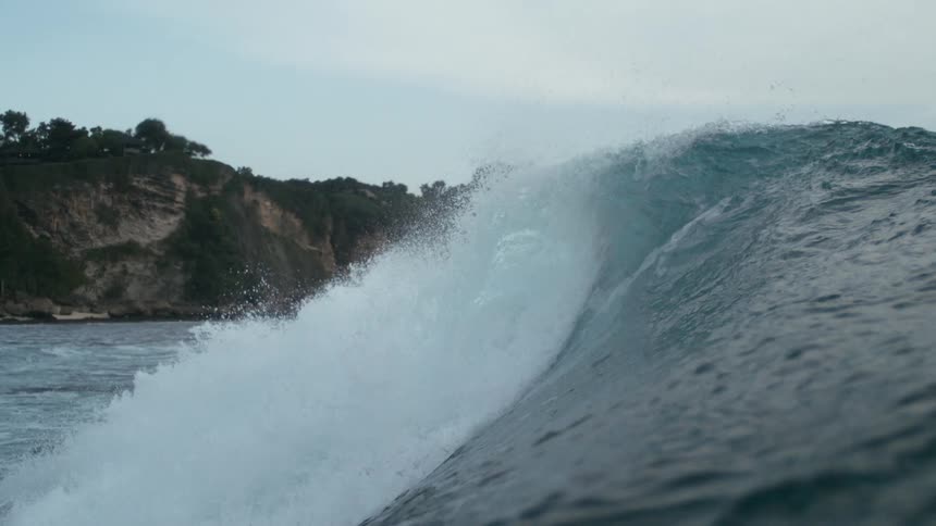 A powerful ocean wave with a breaking crest is seen under a cloudy sky wih a mountain in the background