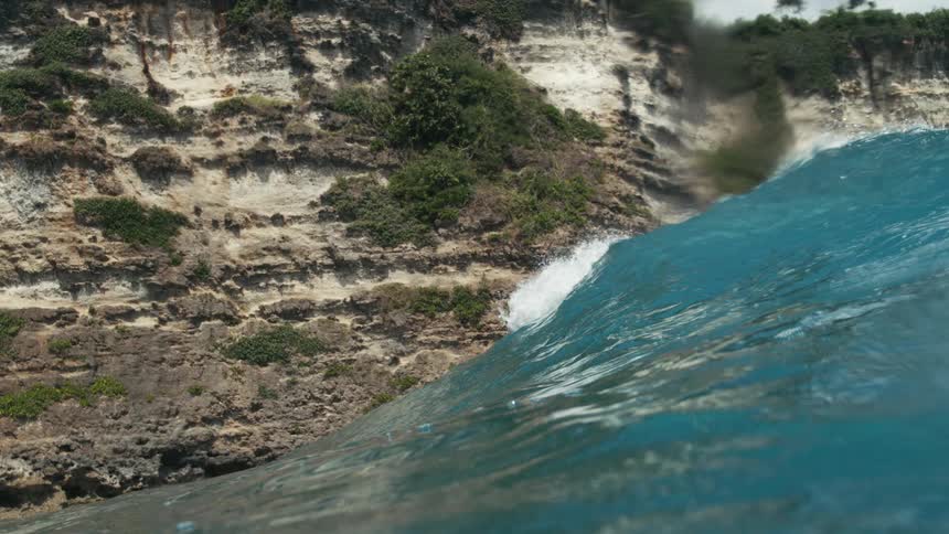 A turquoise ocean wave rises near rugged coastal cliffs