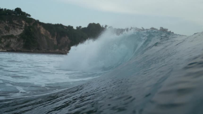 A large ocean wave crashes near a rocky coastline sending spray upward as it breaks in front of vegetationcovered cliffs