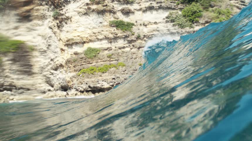 A wave curls and breaks against rocky cliffs topped with green vegetation