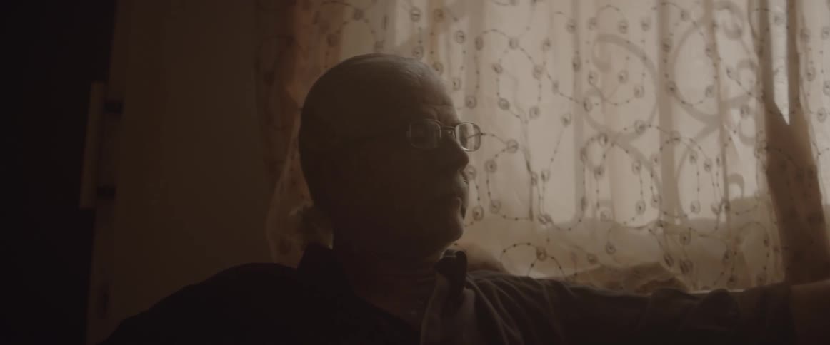 A man sits in front of a curtained window