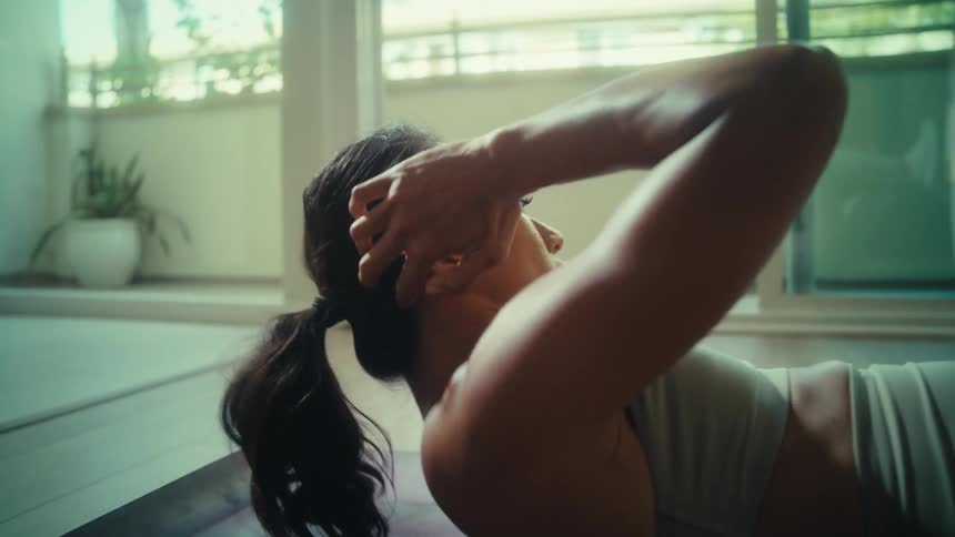 A woman with a ponytail performs a situp on a yoga mat in a sunlit room
