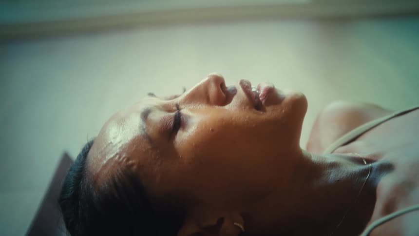 A woman in a light colored tank top lies on a yoga mat on a wooden floor catching her breath after exercise