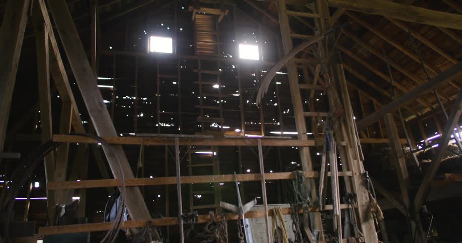 Sunlight filters through two small windows in the roof of an old wooden barn casting light across its aged beams and cluttered interior