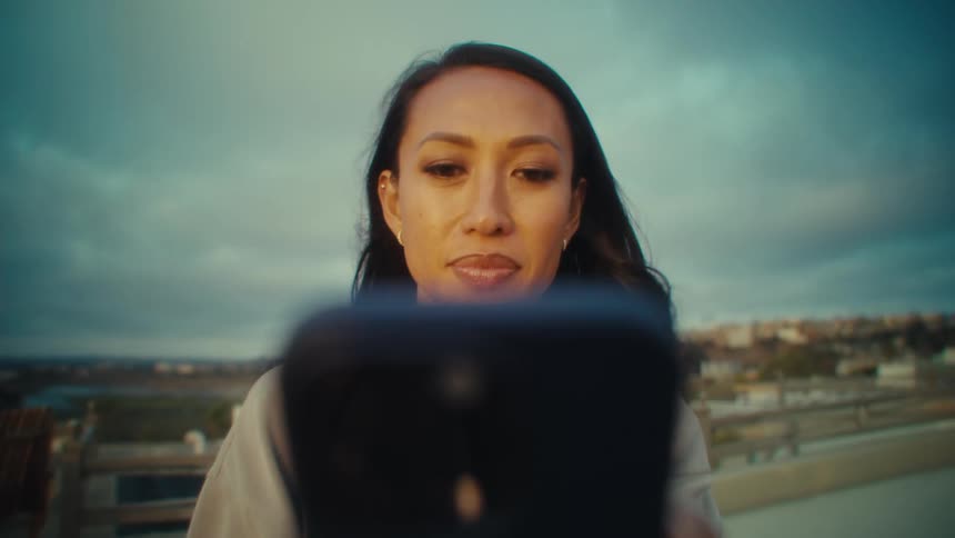A woman holding up a phone for a selfie or video call while cloudy skies and distant buildings stretch beyond the railing