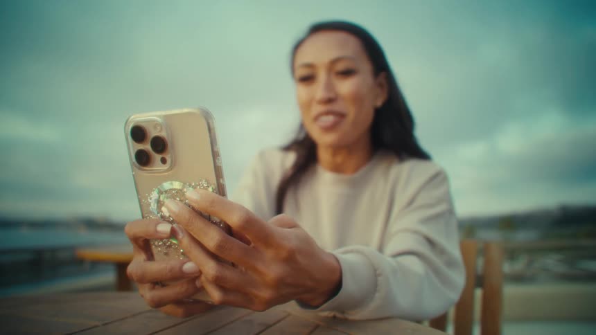 A young woman smiles and speaks on a video call with her phone while sitting at a table outdoors during daytime