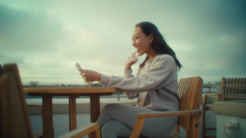 A woman in a light longsleeve shirt and gray pants sits on a wooden deck holding up a phone for a video call while cloudy skies and distant buildings stretch beyond the railing