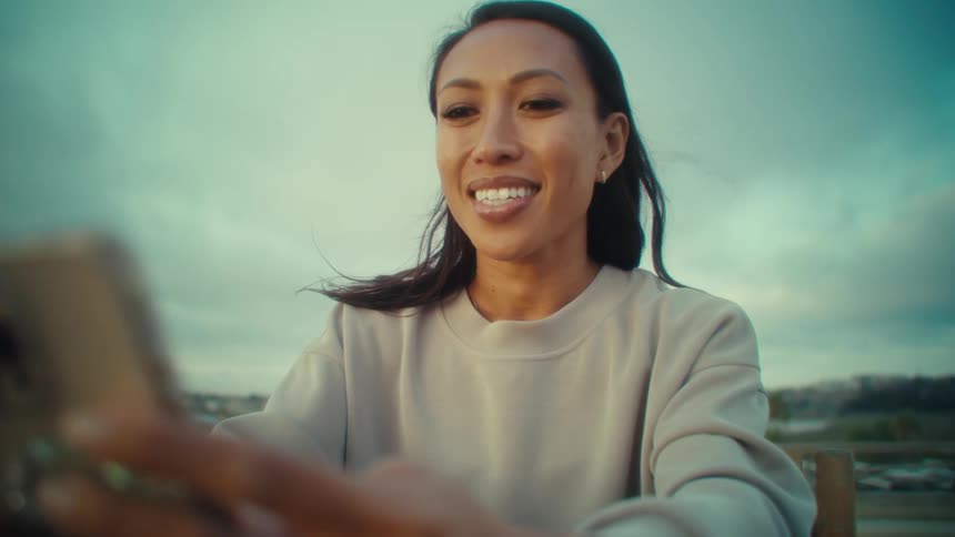 An asian women wearing a lightcolored sweatshirt in an outdoor setting under a cloudy sky looking to her smartphone