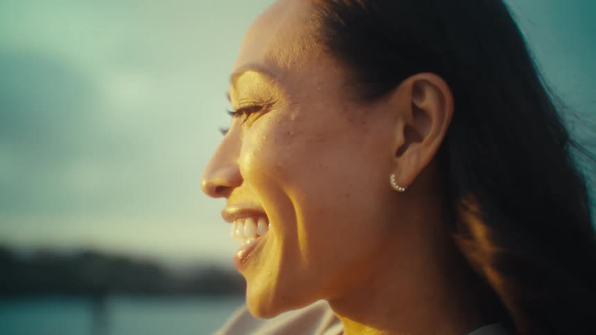 A smiling woman with dark hair illuminated by warm golden hour sunlight against a blurred background of a body of water and sky