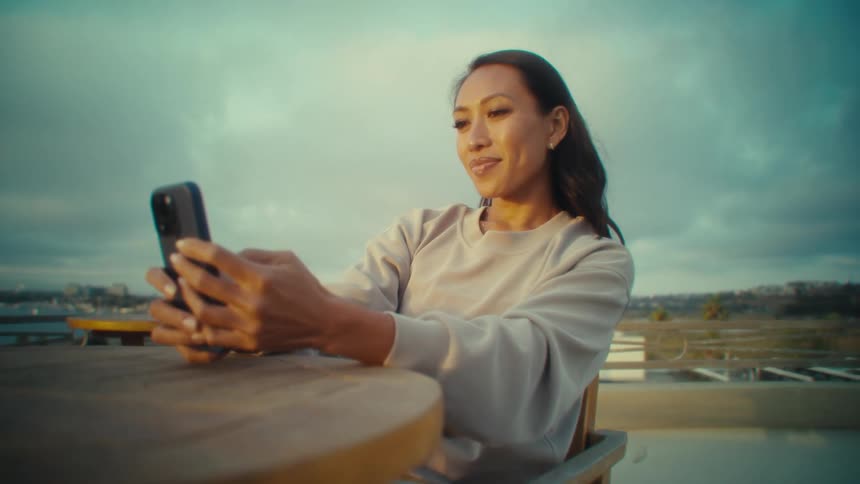 A woman in a light longsleeve shirt and gray pants sits on a wooden deck holding up a phone for a selfie or video call while cloudy skies and distant buildings stretch beyond the railing