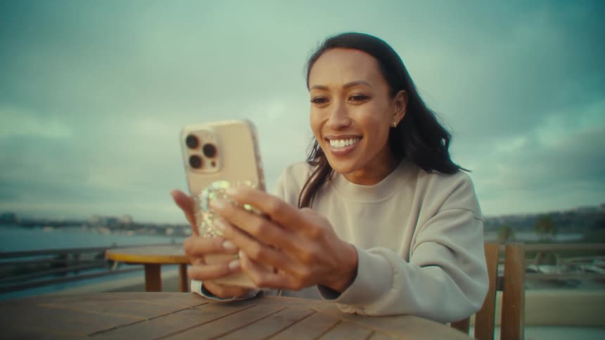 A young woman waves and smiles on a video call with her mobile phone while sitting at a table outdoors during daytime