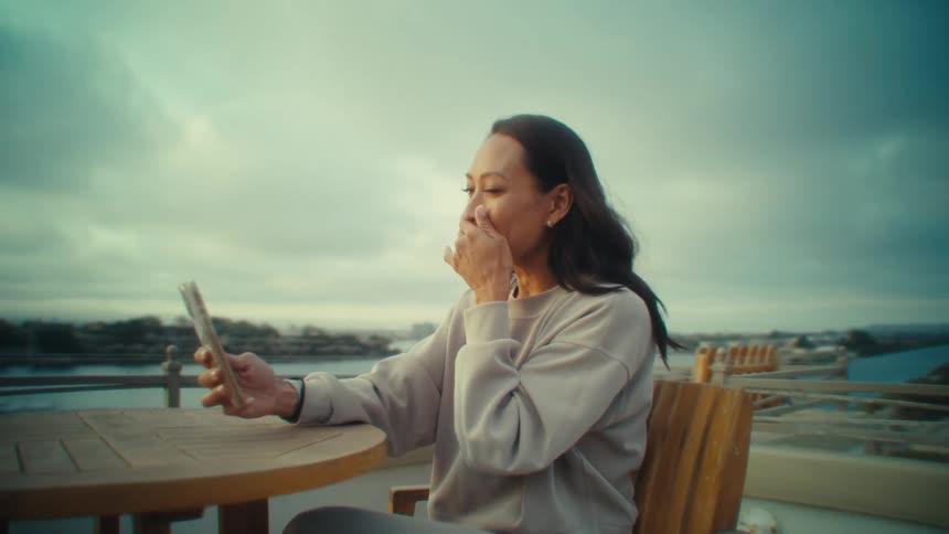 A woman in a light longsleeve shirt and gray pants sits on a wooden deck holding up a phone for a selfie or video call while cloudy skies and distant buildings stretch beyond the railing