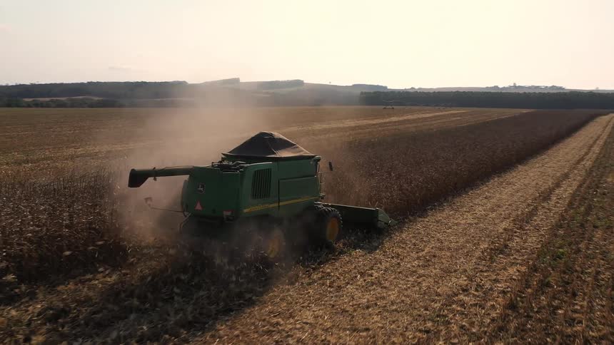A combine harvester moves through a dusty cornfield leaving a clear swath of harvested land under a hazy sky