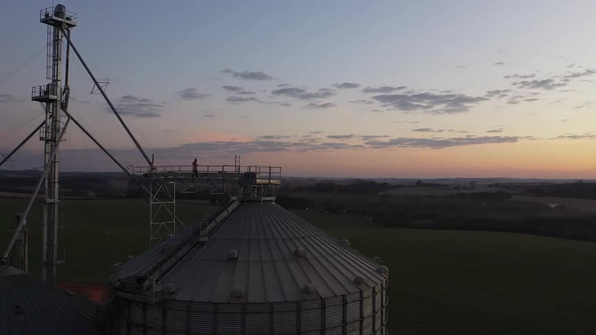 A person stands on top of a grain silo at sunrise overlooking farmland