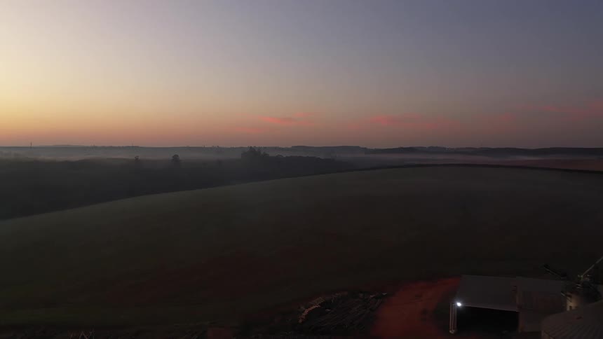 The video provides an aerial perspective of a farm at sunrise or sunset revealing large fields and several farm buildings including multiple tall silos the sky transitions from a bright horizon to darker hues overhead with some mist or fog in the dist