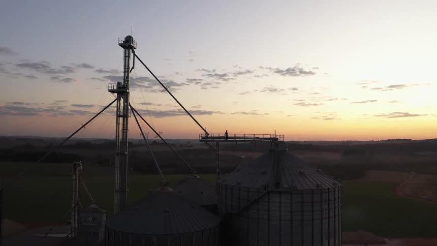The video shows a wide aerial shot of multiple large metallic grain silos and associated farm structures set against a beautiful sunrise or sunset a person can be seen standing on a platform attached to one of the taller silos looking out at the sprawl