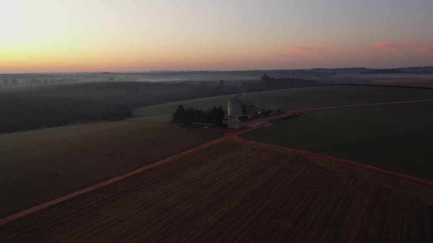 The video captured by a drone showcases a vast expanse of farmland at dusk with the horizon glowing orange and pink in the distance several grain silos stand tall subtly illuminated against the fading light