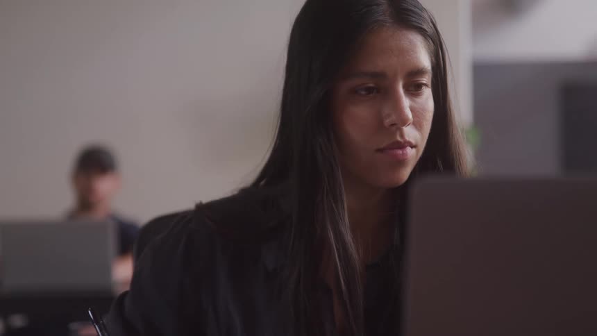 A woman with long dark hair wearing a dark shirt looks down intently at a laptop screen while holding a pen with another person working on a laptop in the blurred background