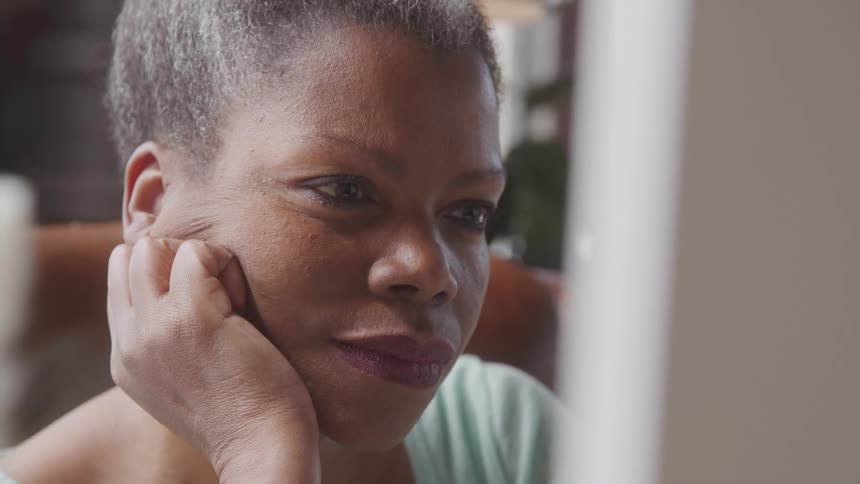 A woman with short grey hair and dark skin rests her chin on her hand looking intently off to the right side of the frame with a serious expression