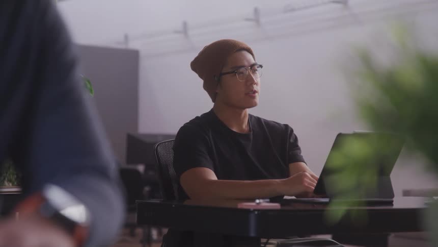 A man with a beanie and glasses raising his hand while sitting at a desk with a tablet
