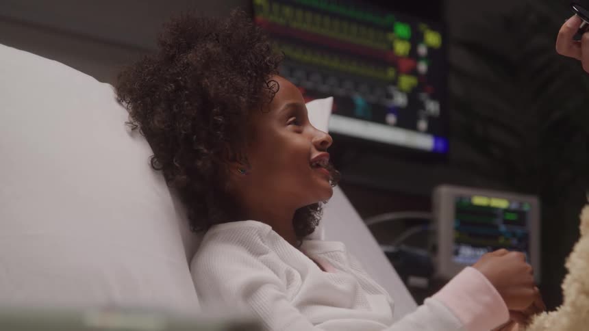 A young girl with dark curly hair lies in a hospital bed looking towards a persons near her with medical equipment in the blurred background